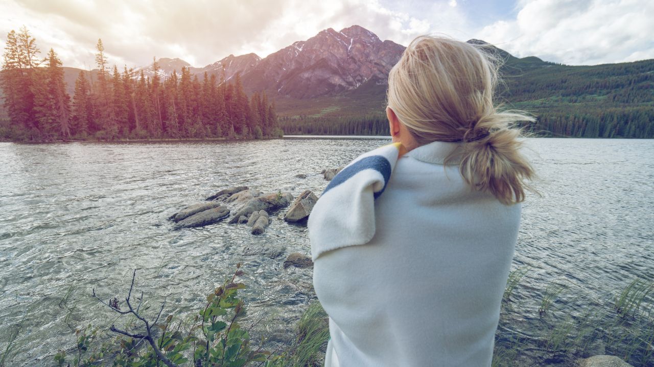 A woman in midlife wrapped in a blanket, standing by a mountain lake and looking toward the horizon, reflecting on her life and what comes next.