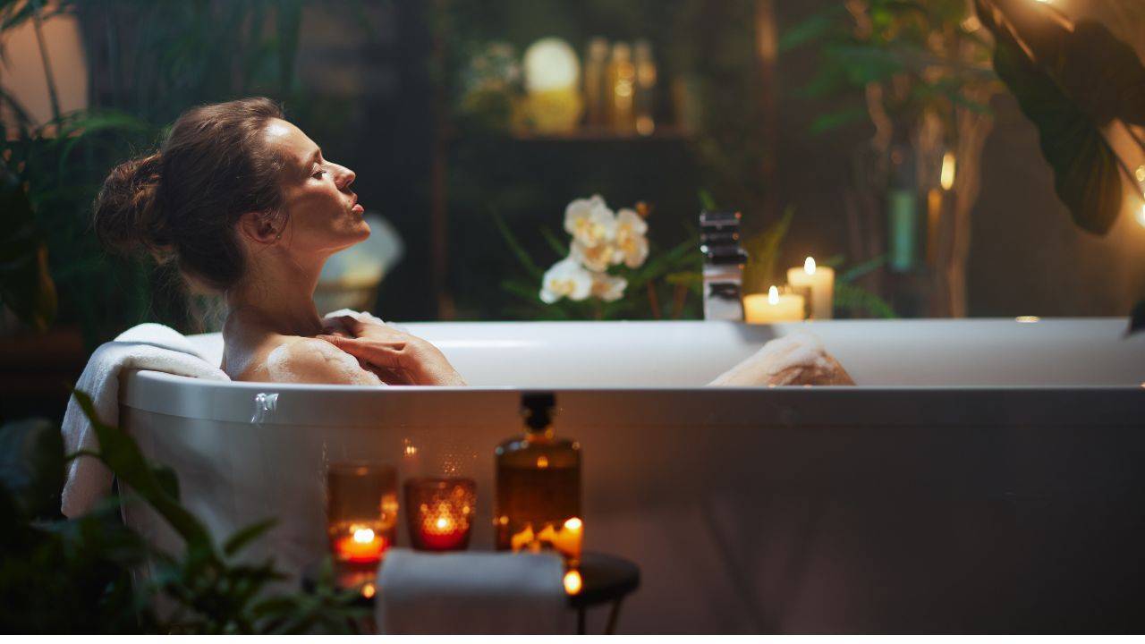 A woman relaxing in a warm bath surrounded by candles and plants, representing peaceful and affordable self-care for women in midlife.