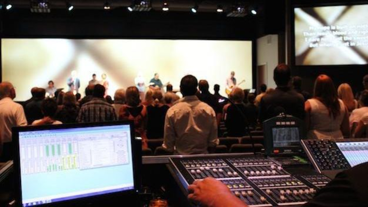 Wide-angle view of a church worship service with a full band on stage, showing a slightly misaligned microphone, out-of-sync lyrics on the LED screen, uneven stage lighting, and a congregation with mixed engagement.