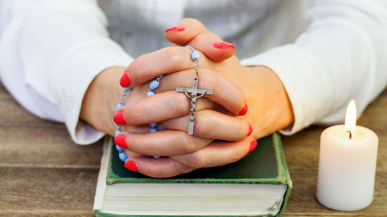 Hands holding a rosary in prayer, resting on a Bible beside a lit candle.