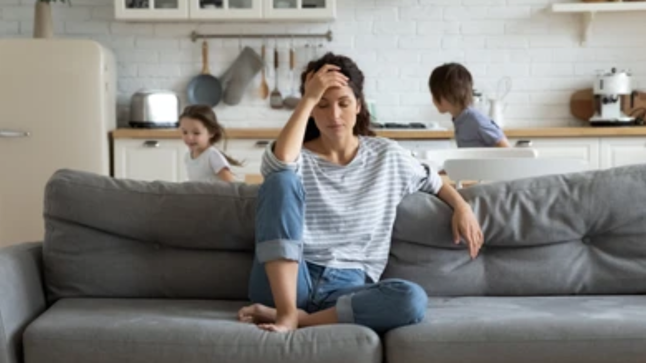 Mom sitting on a couch with her hand on her forehead, looking overwhelmed while young children play in the kitchen behind her.
