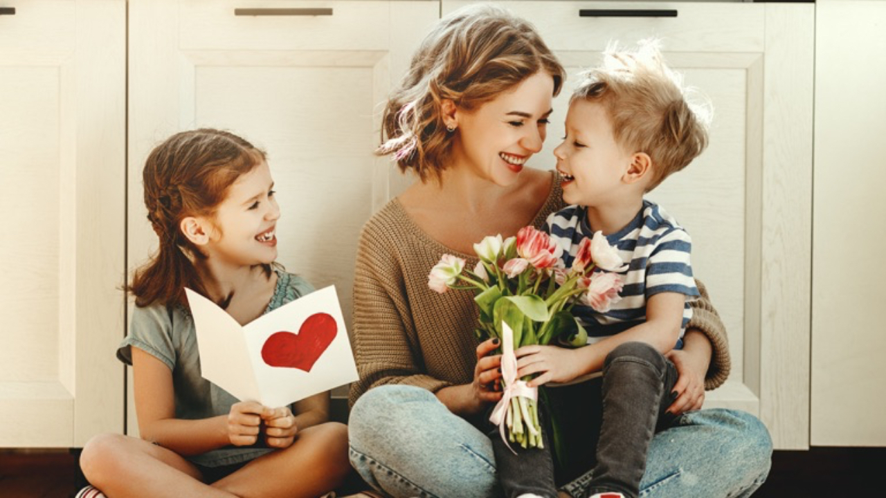 A mother sits on the floor smiling with her two children, who give her flowers and a handmade card with a heart.