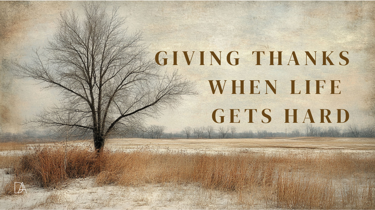 A winter landscape featuring a bare tree standing alone in a field of dried brown grass with light snow on the ground. The sky is muted and overcast, giving the scene a calm, somber tone. On the right side of the image, large serif text reads “Giving Thanks When Life Gets Hard.” A small “DA” logo appears in the bottom left corner.