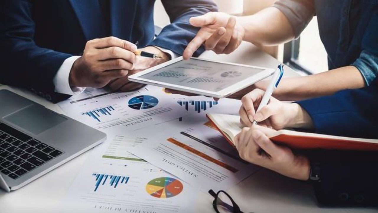 Three business professionals, including a founder or business owner, reviewing financial reports and charts on a tablet and paper, while taking notes during a strategic year-end planning meeting.