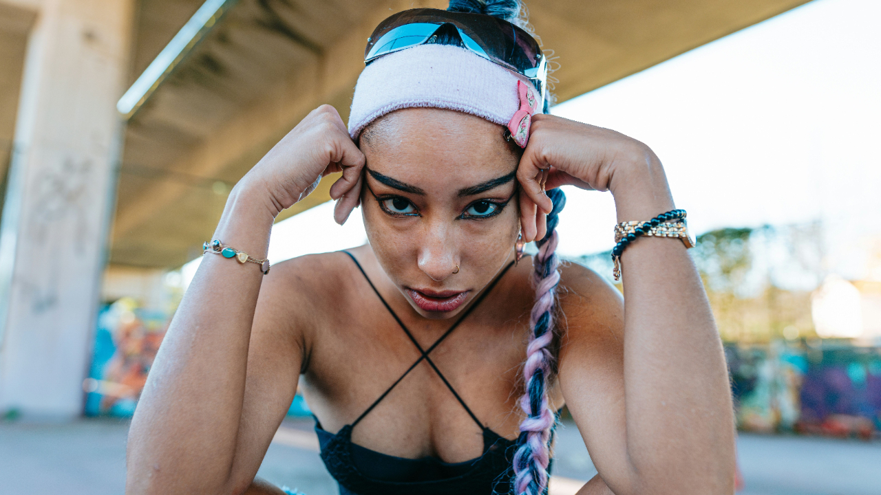 A woman sitting under an overpass, staring directly into the camera with intensity and emotion. Her posture and expression reflect struggle, resilience, and personal transformation.
