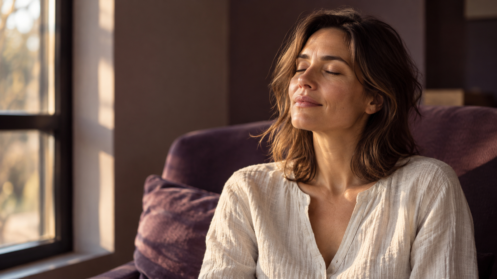 Woman with eyes gently closed beside a sunlit window, reflecting quietly in a purple chair.