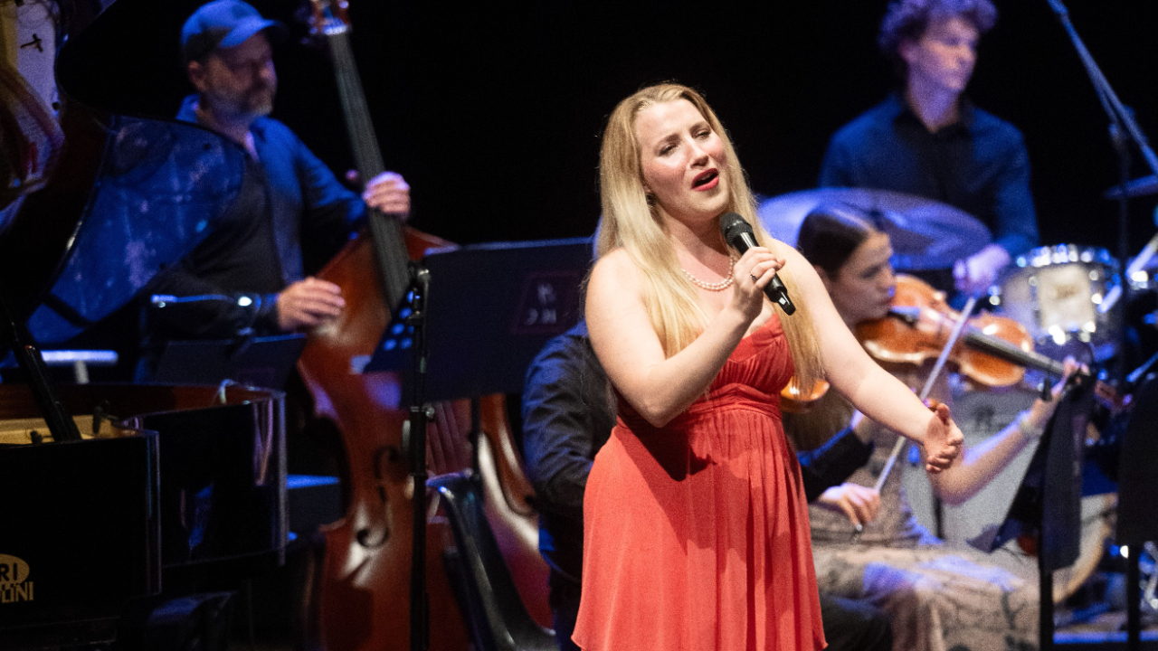 Olivia Kellett, a blonde woman in a red dress, singing on stage with orchestra musicians visible behind her.