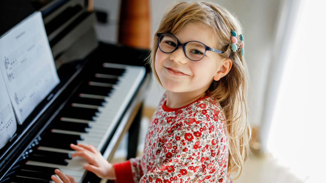 Child wearing glasses looking up and smiling with hands on piano keyboard