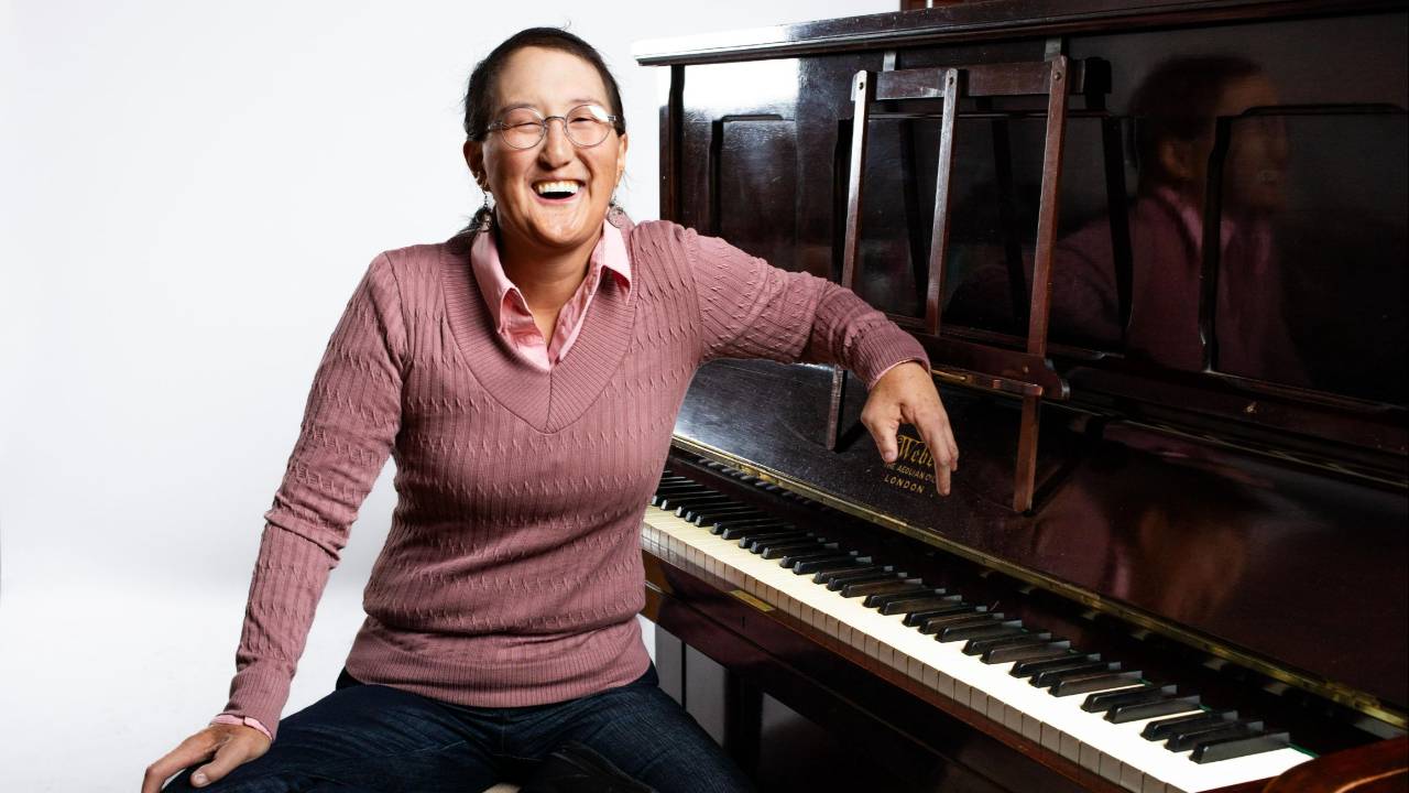 Piano teacher, Camille Hatton, is a Korean American person in her late 30s. She is seated comfortably at an upright piano bench wearing small round glasses, thin dangling earrings, a pink collared shirt and sweater, and a big smile.