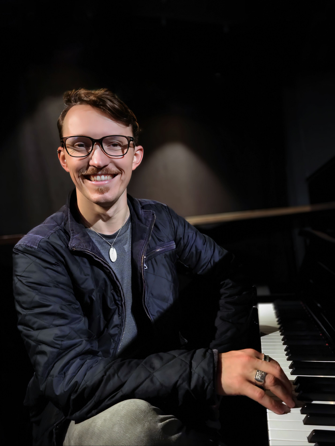 Chase Chandler, a smiling man with short brown hair and glasses, seated at a piano with his right hand resting on the keys.