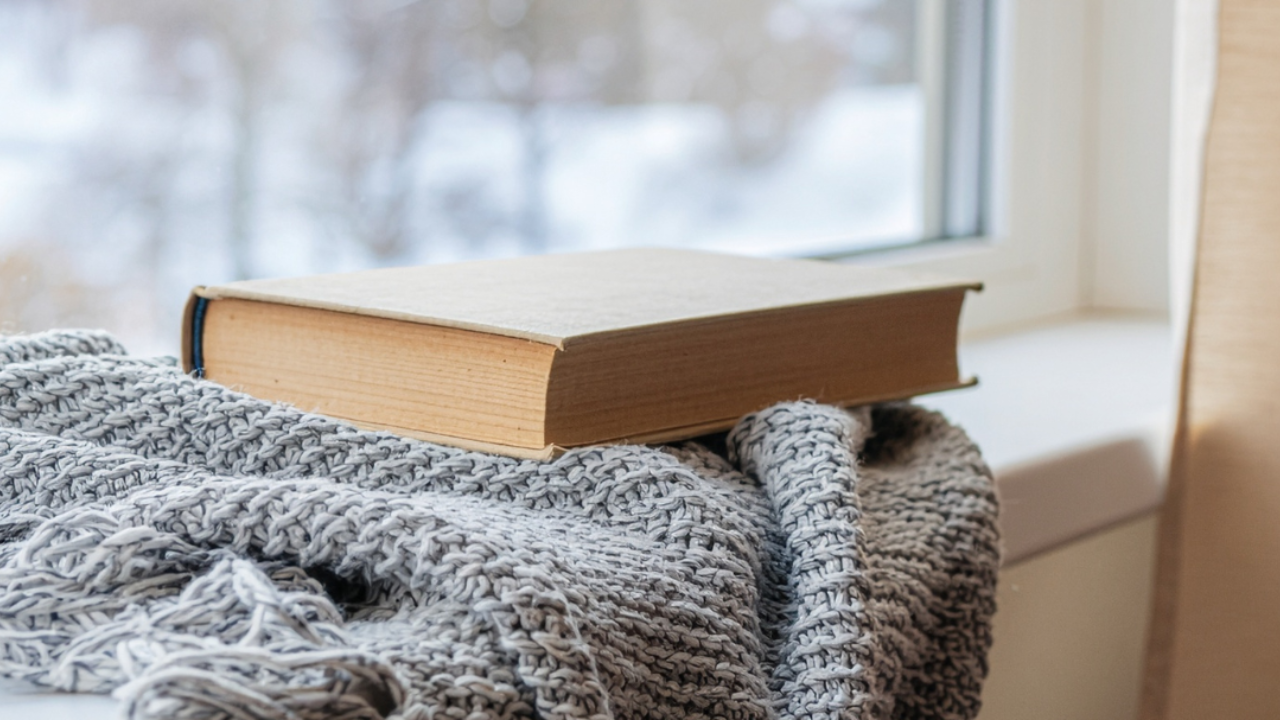 A closed book resting on a knit blanket by a snowy winter window, evoking a quiet, gentle January moment.