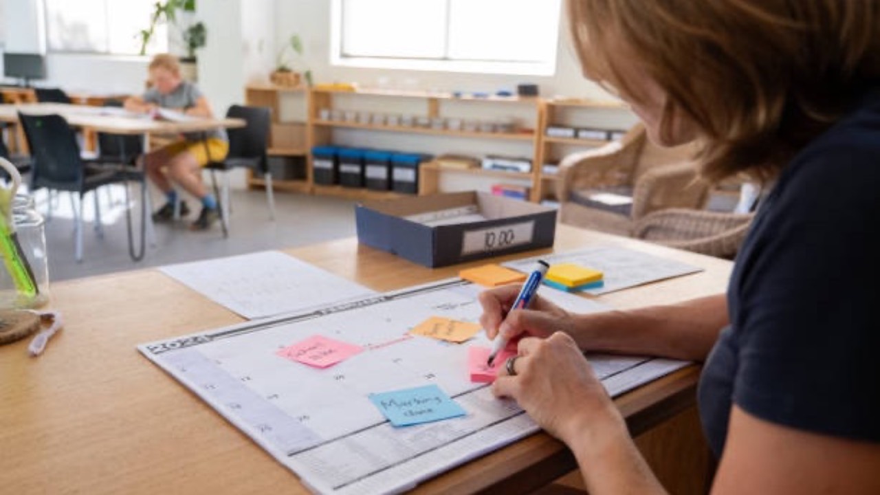 Childcare center director standing in an empty classroom at sunset holding a notebook and calendar, looking thoughtful while paperwork piles sit on a table in front of her.