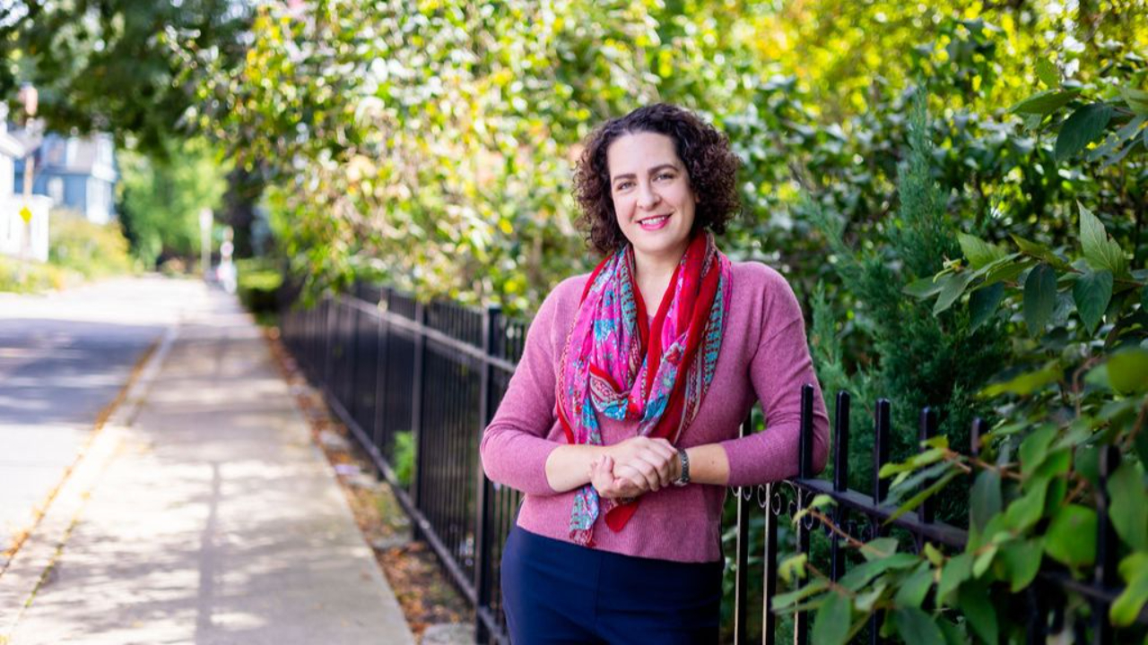 Erica Mattison standing outdoors by a fence and greenery on a tree-lined sidewalk.