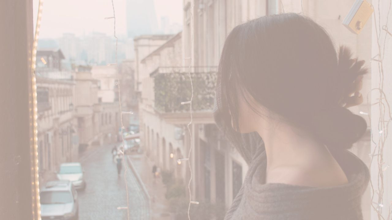 Midlife woman standing by a window, softly reflecting as she looks out over a quiet city street, representing stress and emotional shifts during perimenopause.