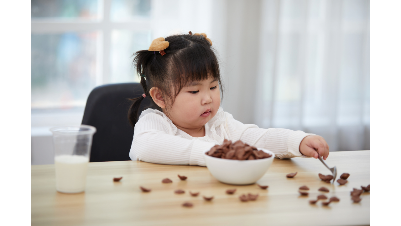 A young child concentrates while using a spoon to scoop cereal from a bowl at a table, with a few spills visible around them.