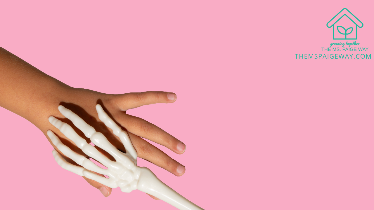 A child’s hand resting against a white plastic skeleton hand on a soft pink background, symbolizing connection and inclusion during Halloween.