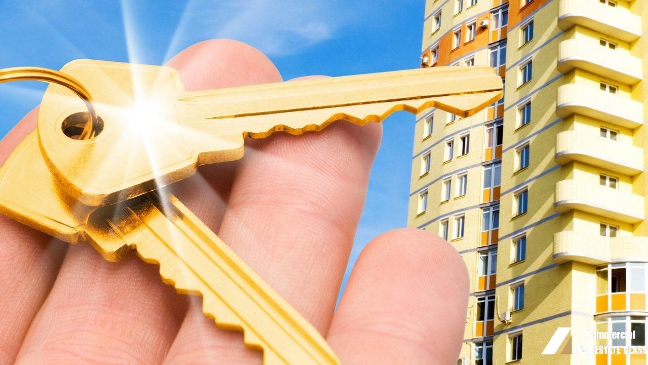 Hand holding gold keys in front of a commercial apartment building representing property ownership and real estate value