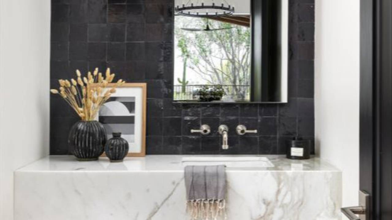 Modern bathroom vanity with a marble sink and dark Zellige tile feature wall, styled with black pendant lighting, brass tapware, and decorative accents.