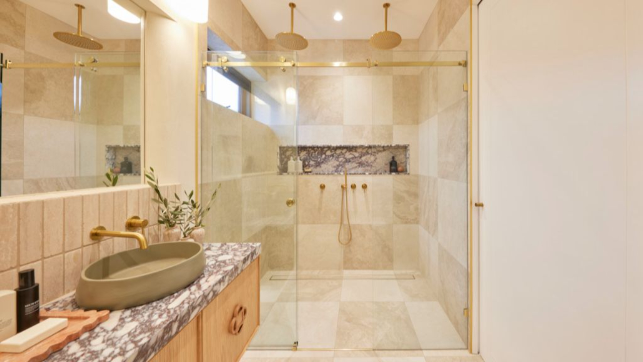 Luxurious main ensuite bathroom from The Block 2024 featuring a walk-in double shower with brass fixtures, travertine chequerboard tiles, olive green basin, and marble vanity top.