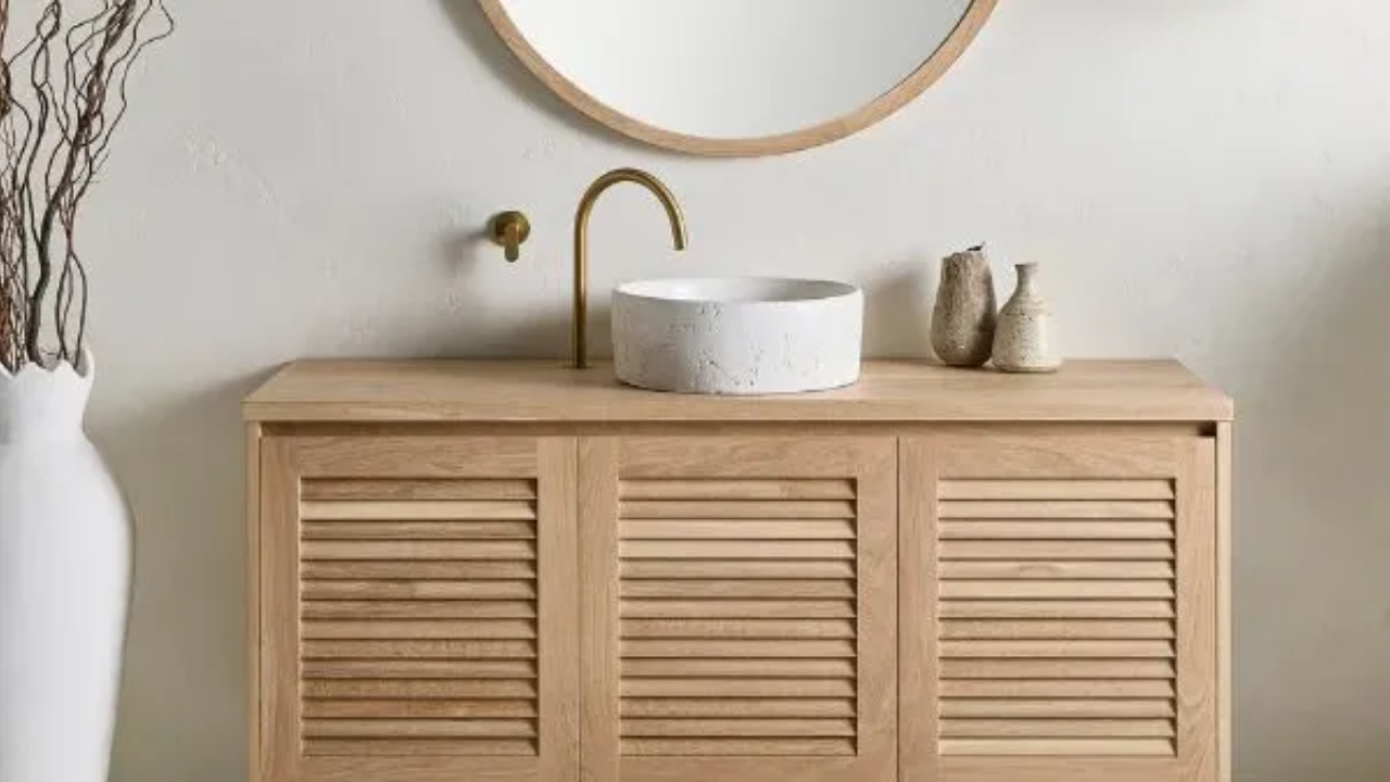 Coastal-style bathroom vanity in natural oak with louvre doors, round mirror, white textured basin, and brass wall-mounted tapware against a soft neutral wall.