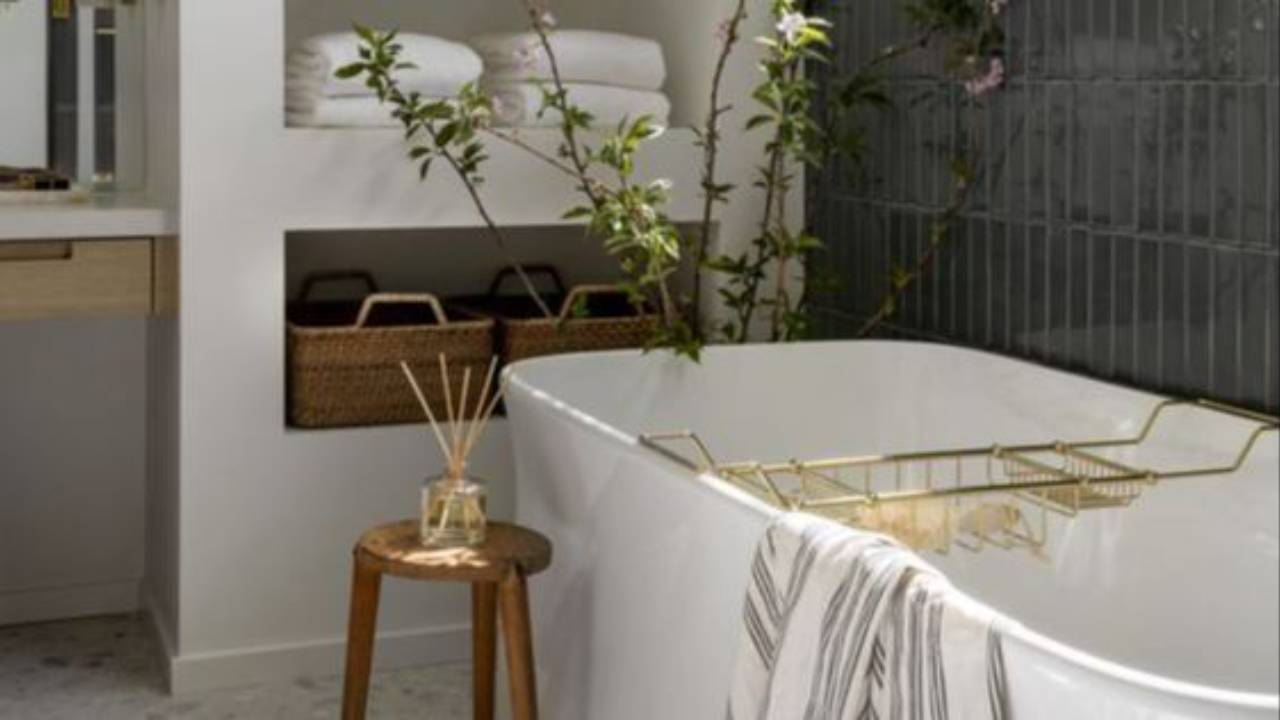 Modern bathroom with back-to-wall freestanding bath, vertical green tiles, open shelving, brass accessories, and large feature window providing natural light.