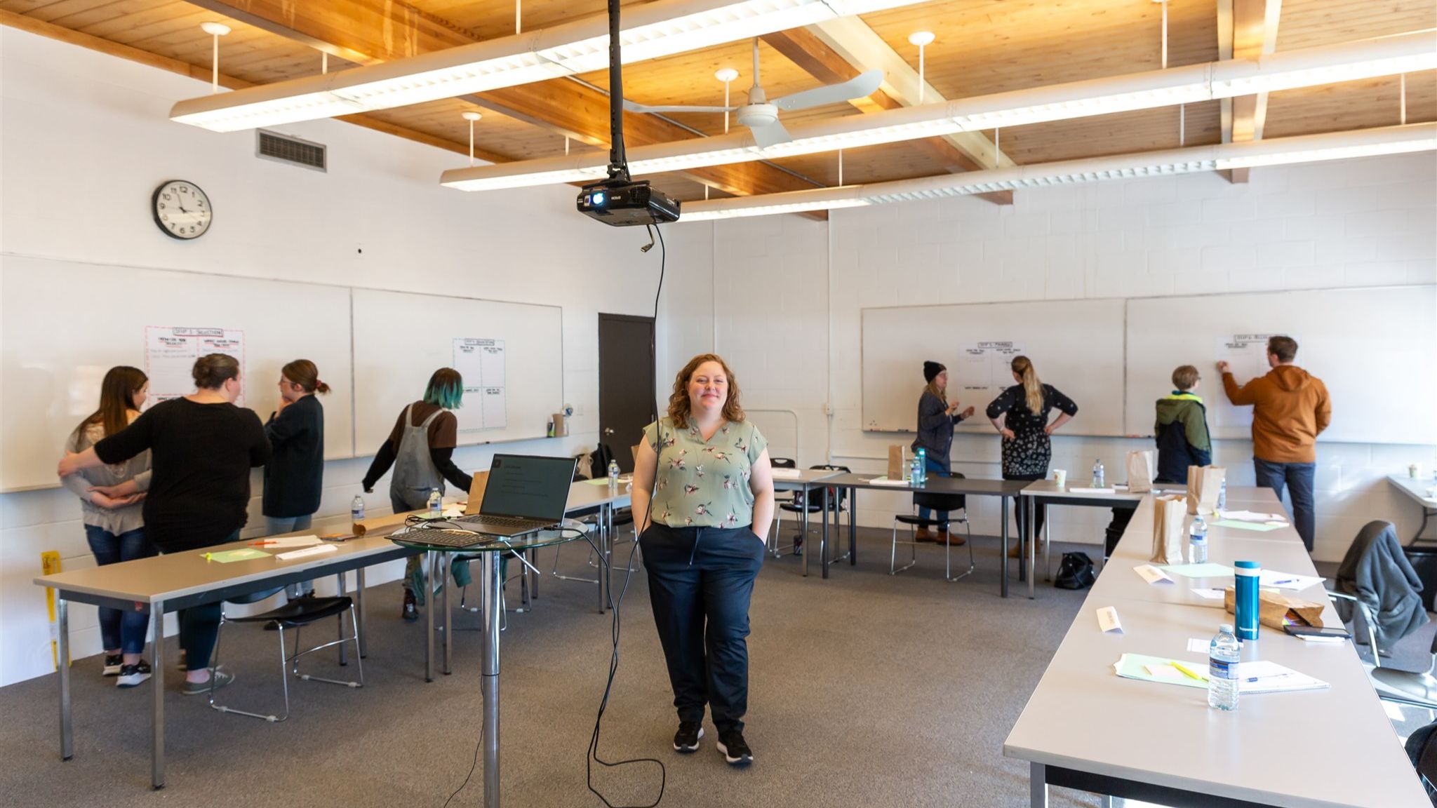 Group of adults standing around tables in a bright workshop space, participating in a hands-on strategy session with notebooks and materials spread out.