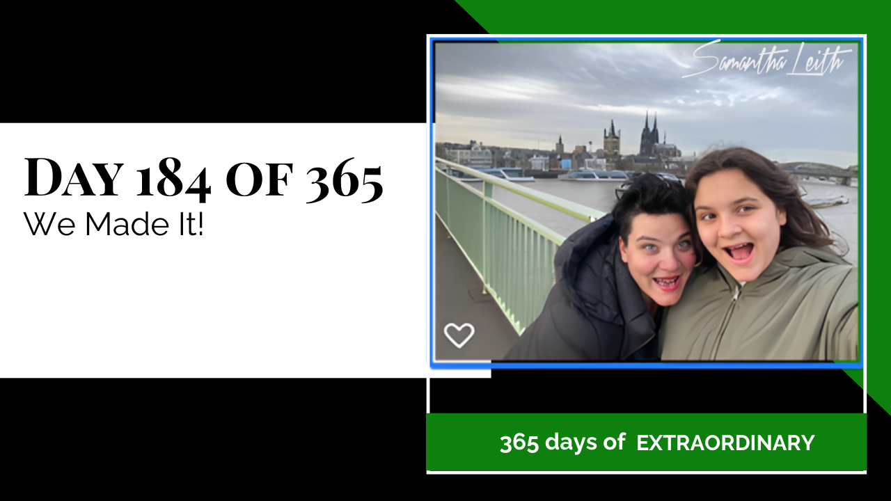 A close-up, outdoor selfie of two women bundled in winter coats, smiling excitedly on a bridge with the Cologne Cathedral and city skyline visible, representing successful international travel, arrival at a new destination, and the completion of a long journey.