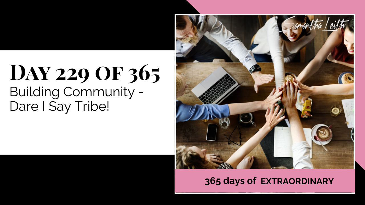  A photographic post featuring an overhead shot of a diverse group of seven people gathered around a wooden table, stacking their hands together in the center in a show of teamwork or unity. A laptop, mugs, and papers are scattered on the table. The image is split with a title card on the left side that reads: "DAY 229 OF 365 Building Community - Dare I Say Tribe!". Below the photo, a bright pink banner reads: "365 DAYS OF EXTRAORDINARY". A stylized signature in the top right corner says "Samantha Leith."