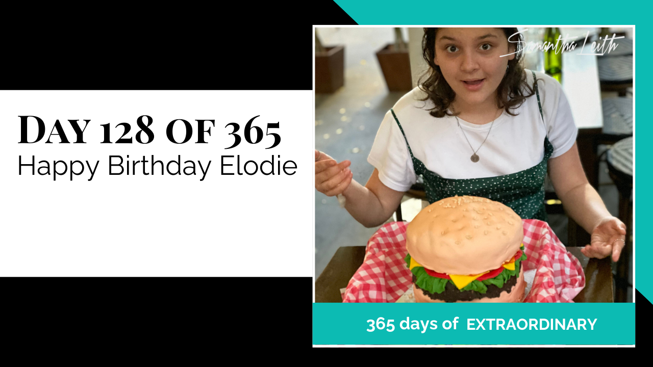 Blog image for Day 128 of Sam Leith's '365 Days of Extraordinary' series, showing a young woman (Elodie) smiling while seated at a table with a large, realistic cake shaped like a giant hamburger wrapped in a checkered napkin, celebrating her birthday.
