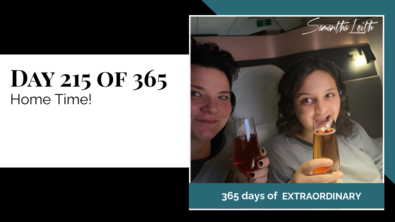 A photographic post featuring two people smiling and clinking champagne flutes while sitting next to each other in what appears to be a premium airline cabin. The person on the left has short, dark hair and is holding a glass of dark pink liquid. The person on the right has long, dark, curly hair and is holding a glass of lighter, amber-colored liquid with a piece of fruit floating in it. The image is split with a title card on the left side that reads: "DAY 215 OF 365 Home Time!" Below the photo, a banner reads: "365 DAYS OF EXTRAORDINARY". A stylized signature in the top right corner says "Samantha Leith.