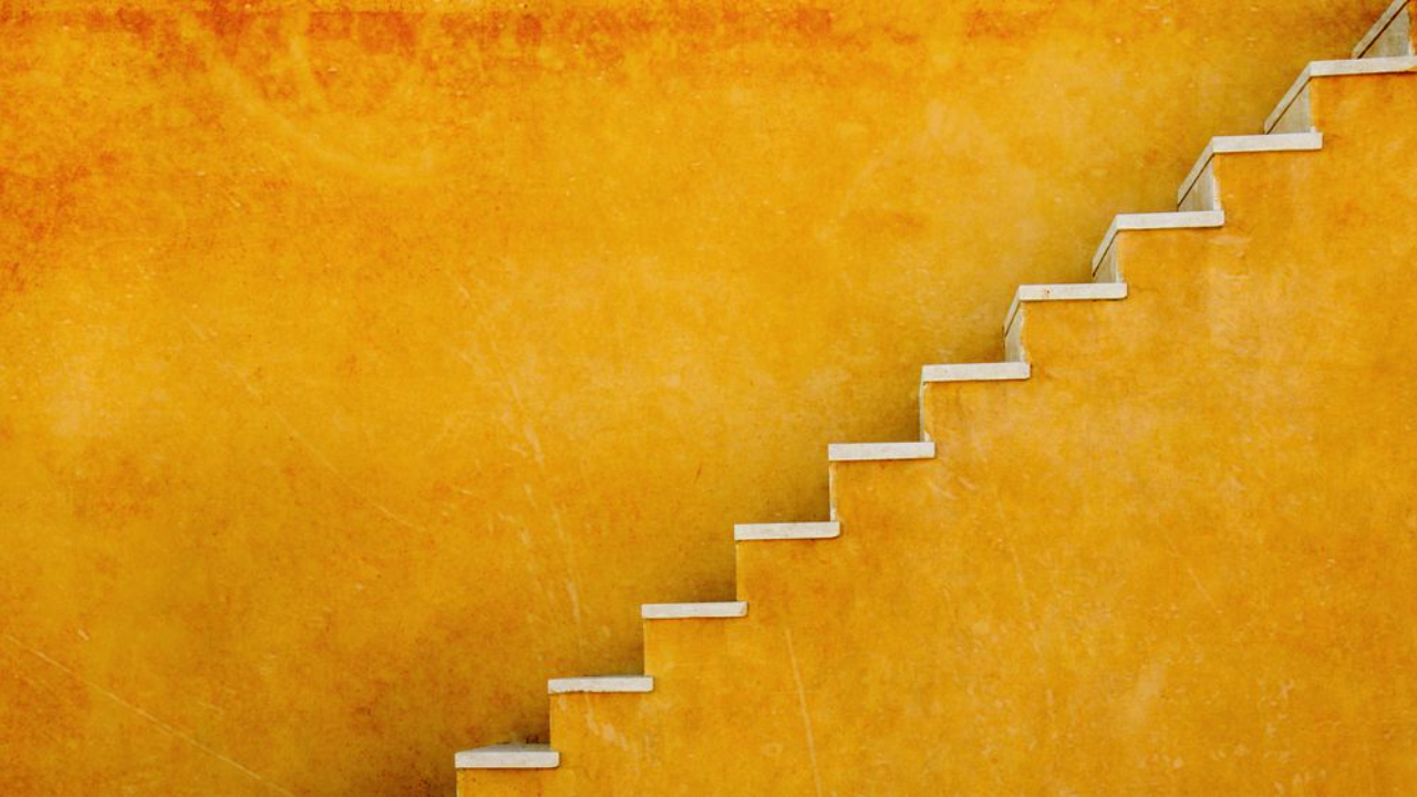 Blog image for the 'Keep it Simple' concept, showing a minimal set of white stairs ascending diagonally against a textured, rustic orange or yellow wall, symbolizing simplifying the journey or taking one step at a time.