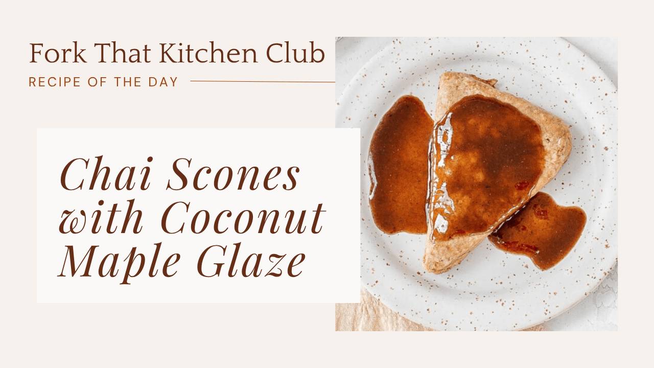A triangular chai scone with coconut maple glaze drizzled on top, placed on a white plate with visible speckles. The glaze pools around the scone, and a gold spoon rests beside the plate on a white marble surface with a cooling rack visible in the background.