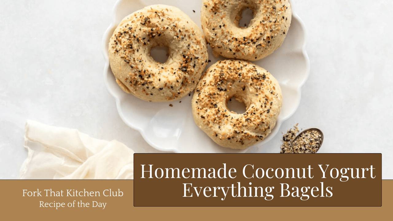 Overhead view of three golden everything bagels arranged on white plate on a white surface with a light ivory linen fabric napkin draped on left side
