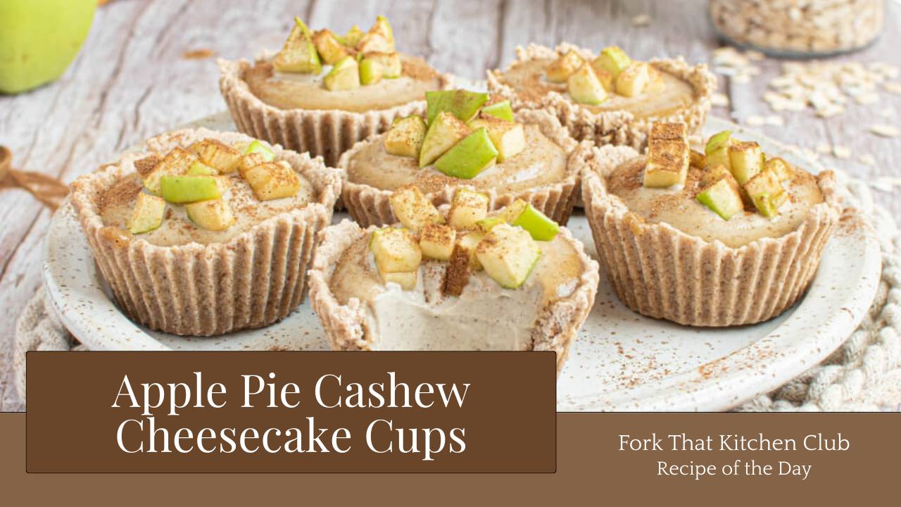 Six cashew cheesecake cups in paper liners arranged on white plate resting on a light wooden surface. Each cup shows a creamy filling topped with diced apple and cinnamon. 