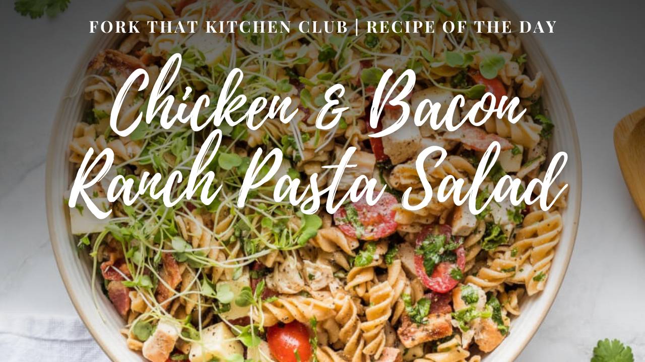 Large white serving bowl of rotini pasta salad with grilled chicken, crispy bacon, cherry tomatoes, and cheddar cheese topped with fresh microgreens on light surface