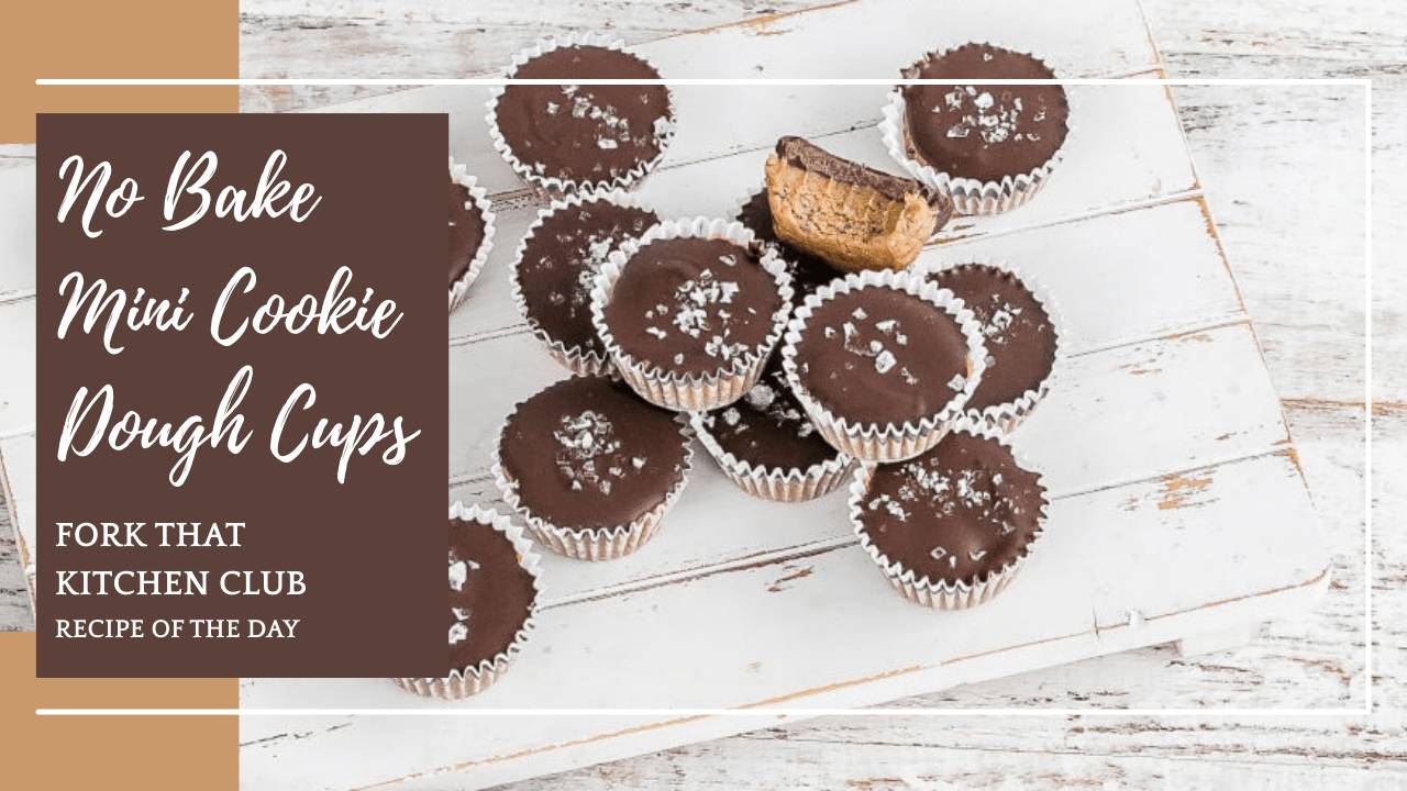 Mini cookie dough cups covered in dark chocolate and topped with flaky sea salt arranged on white surface, several cups scattered across frame with paper muffin liners visible. One cookie dough cup is split in half to highlight the soft cookie dough base