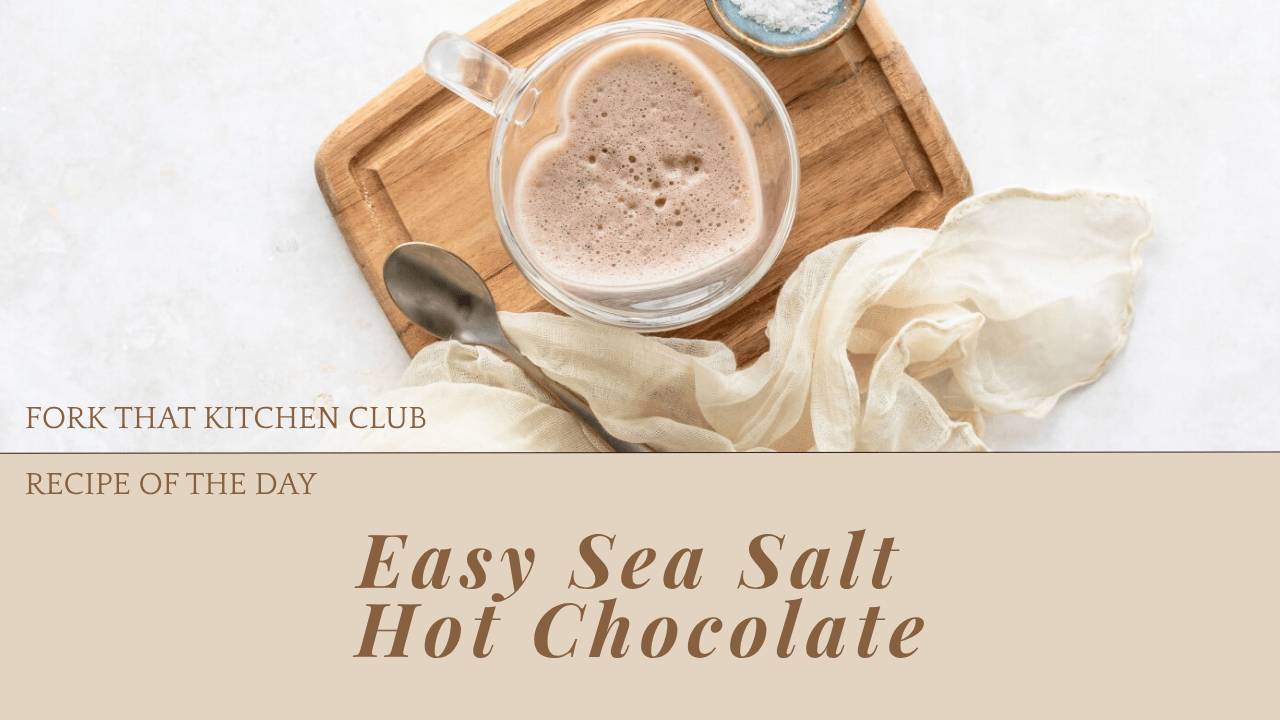 A clear glass mug filled with frothy sea salt hot chocolate sits on a rustic wooden board. The drink has a foamy top dusted with spices. A vintage spoon rests on the board next to the mug, alongside a small bowl of blue sea salt. A cream-colored linen napkin drapes softly in the background, creating a cozy, inviting scene