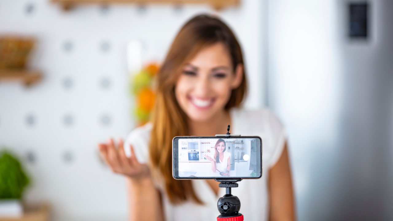 Smiling woman recording a video on a smartphone mounted on a tripod, speaking to the camera in a bright kitchen setting.