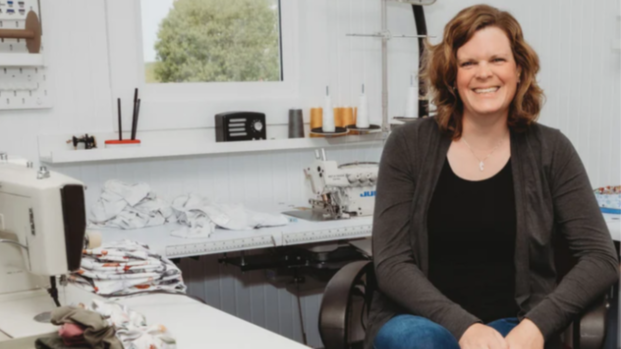 A woman smiling while seated in a bright sewing workspace, surrounded by fabric, folded garments, and industrial sewing machines.