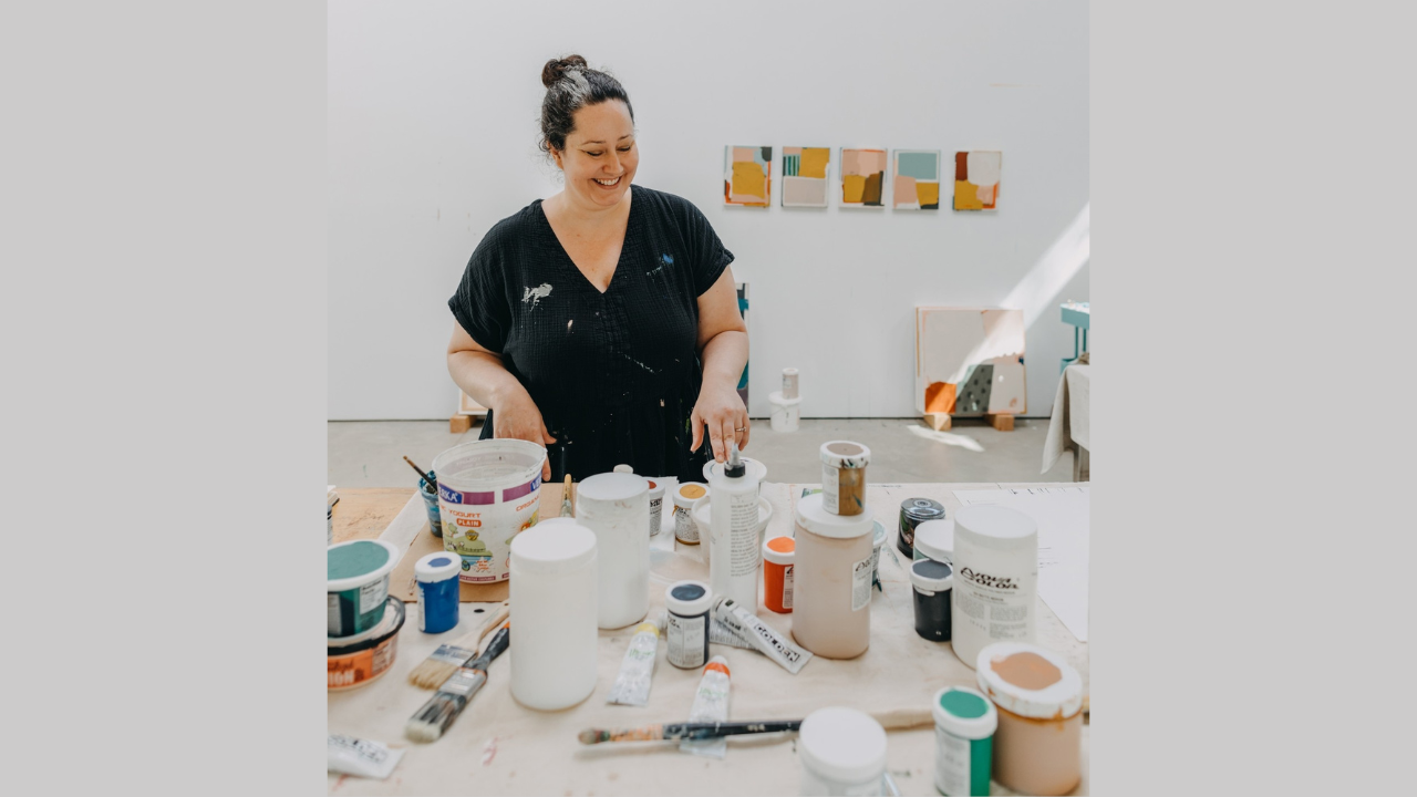 Woman in a black v-neck shirt working at a craft table surrounded by paint containers, brushes, and supplies in a bright studio space with artwork on the walls.