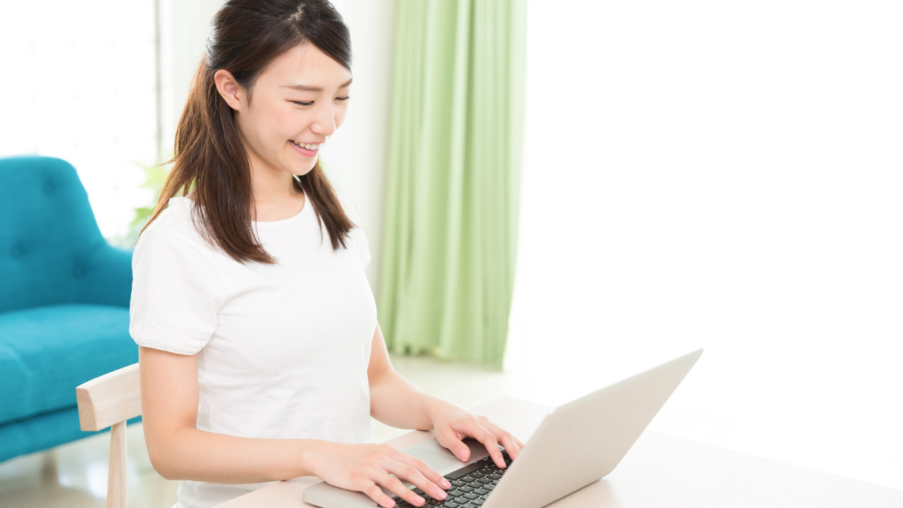 A smiling woman sits at a table typing on a laptop in a bright room, with a blue chair and green curtains in the background.