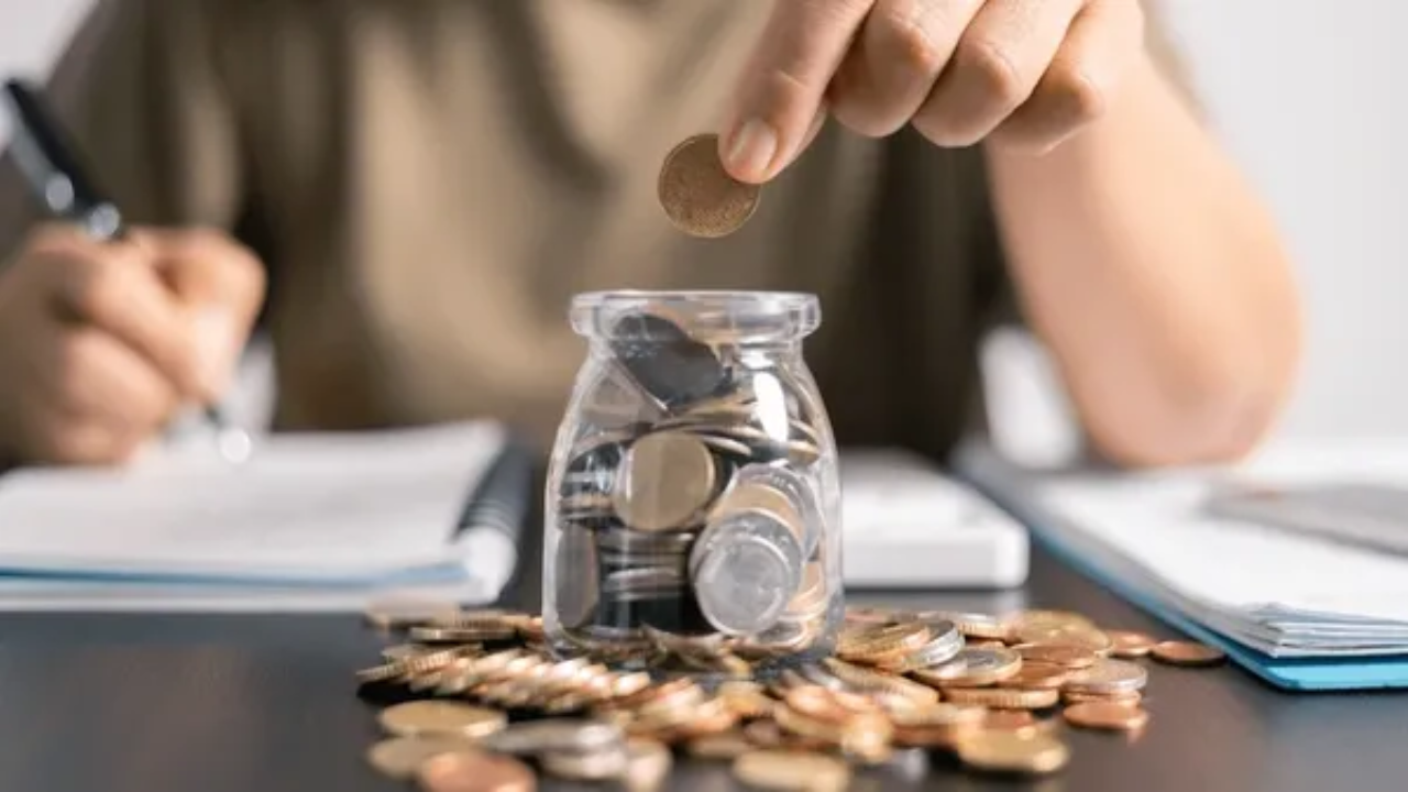 A person placing a coin into a small glass jar filled with money, with scattered coins on the table and budgeting notes in the background.