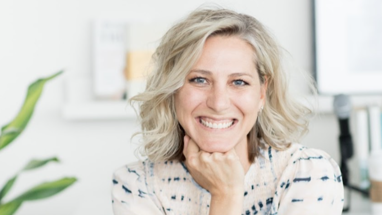 A smiling woman with blonde hair rests her chin on her hand in a bright, modern office setting.
