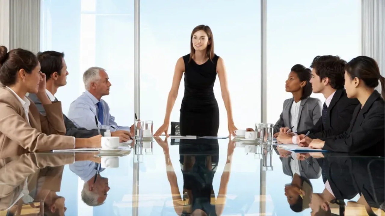 A woman stands confidently at the head of a conference table, leading a meeting with a group of business professionals seated around her in a modern boardroom.