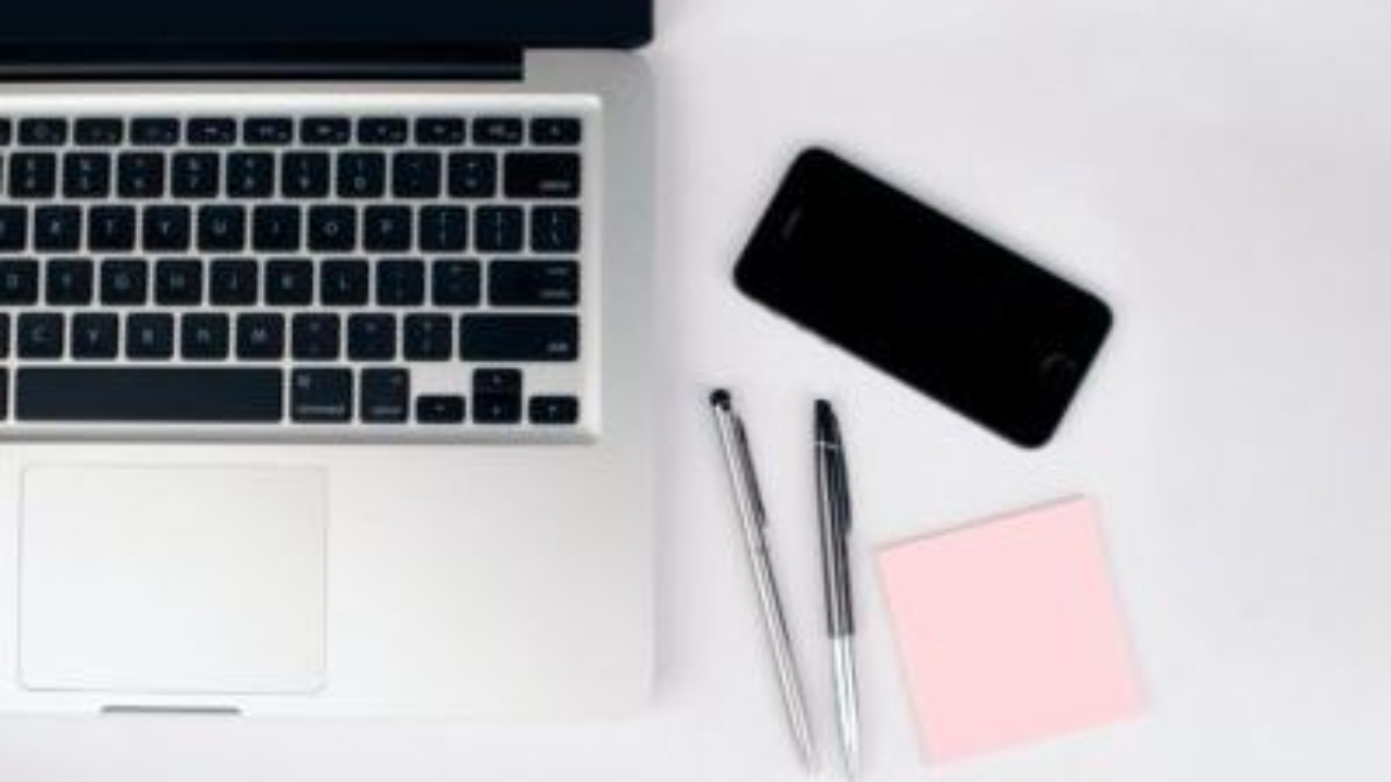 Flat lay of a laptop, smartphone, pens, and pink sticky note on a white desk