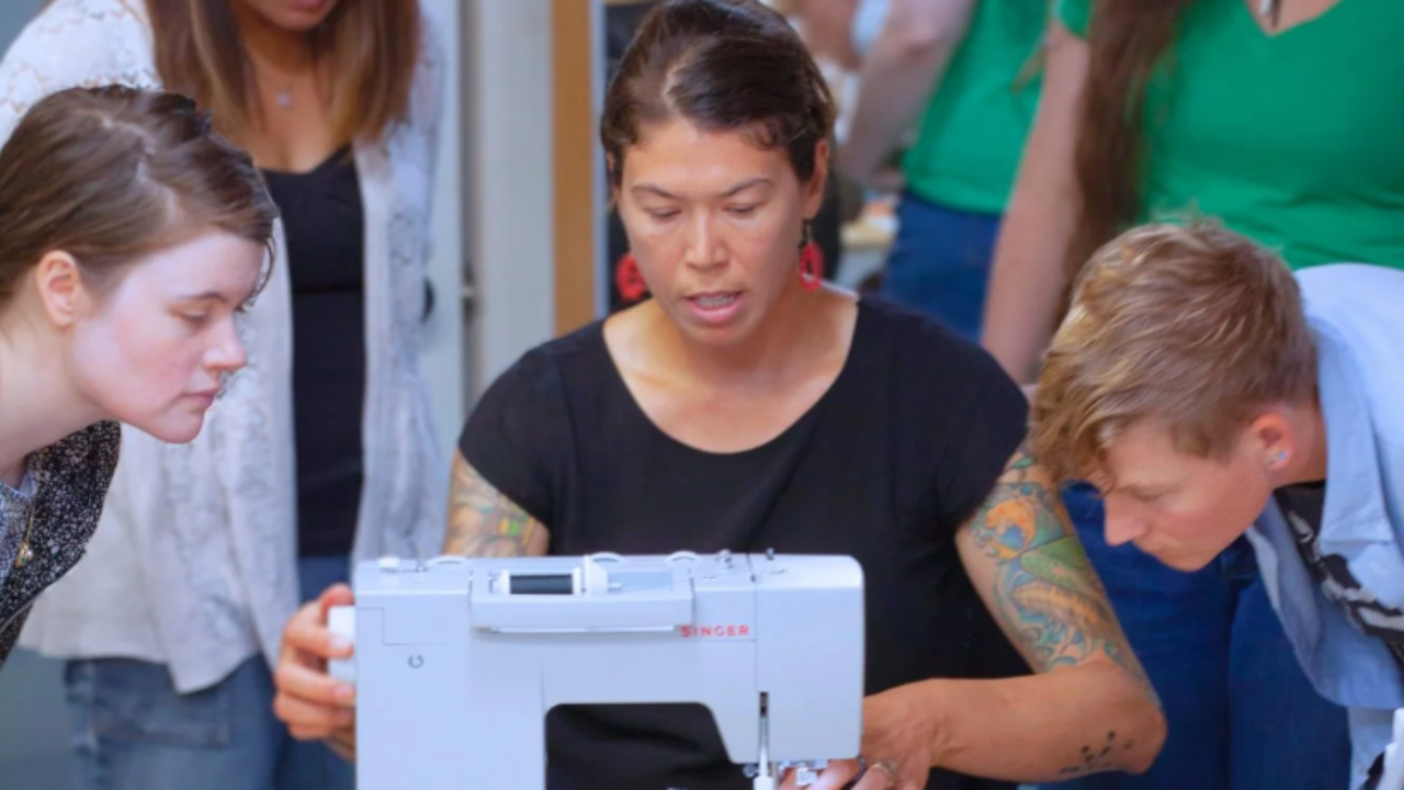 A woman teaching a small group how to use a sewing machine, with students closely watching her demonstrate the technique.