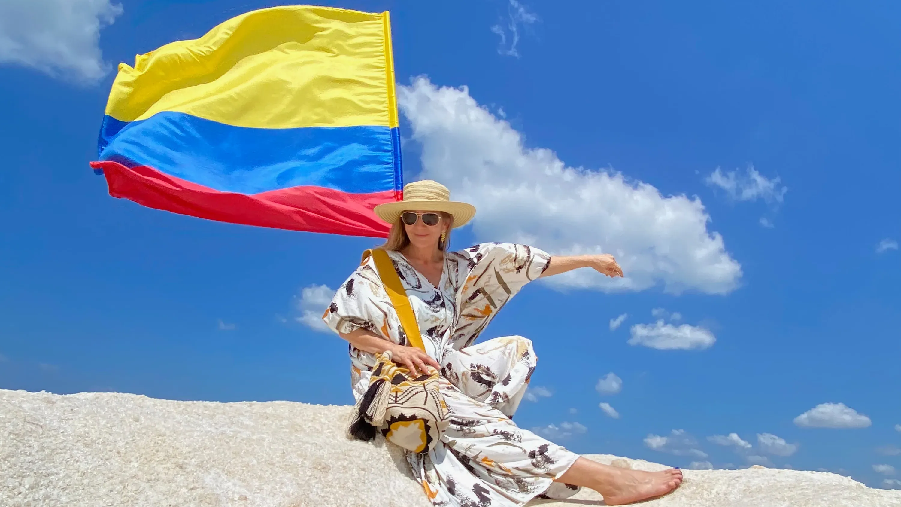 Woman sitting on a white rock under a bright blue sky, wearing a patterned dress and sun hat, holding a handmade woven bag with the Colombian flag waving behind her.