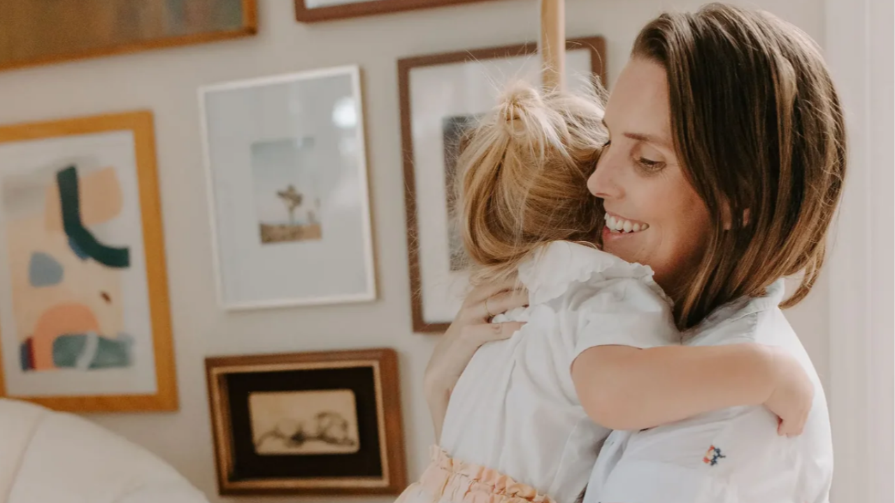 A woman smiling while holding and hugging a young child, standing in a softly lit room with a gallery wall of framed artwork behind them.