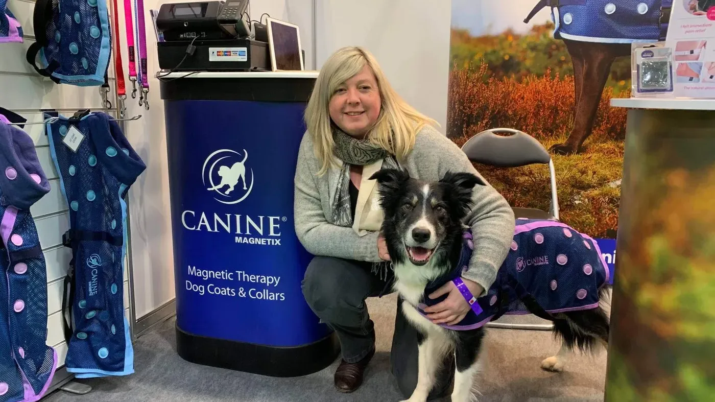 Woman kneeling beside a dog wearing a magnetic therapy coat at a Canine Magnetix booth displaying dog coats and collars.