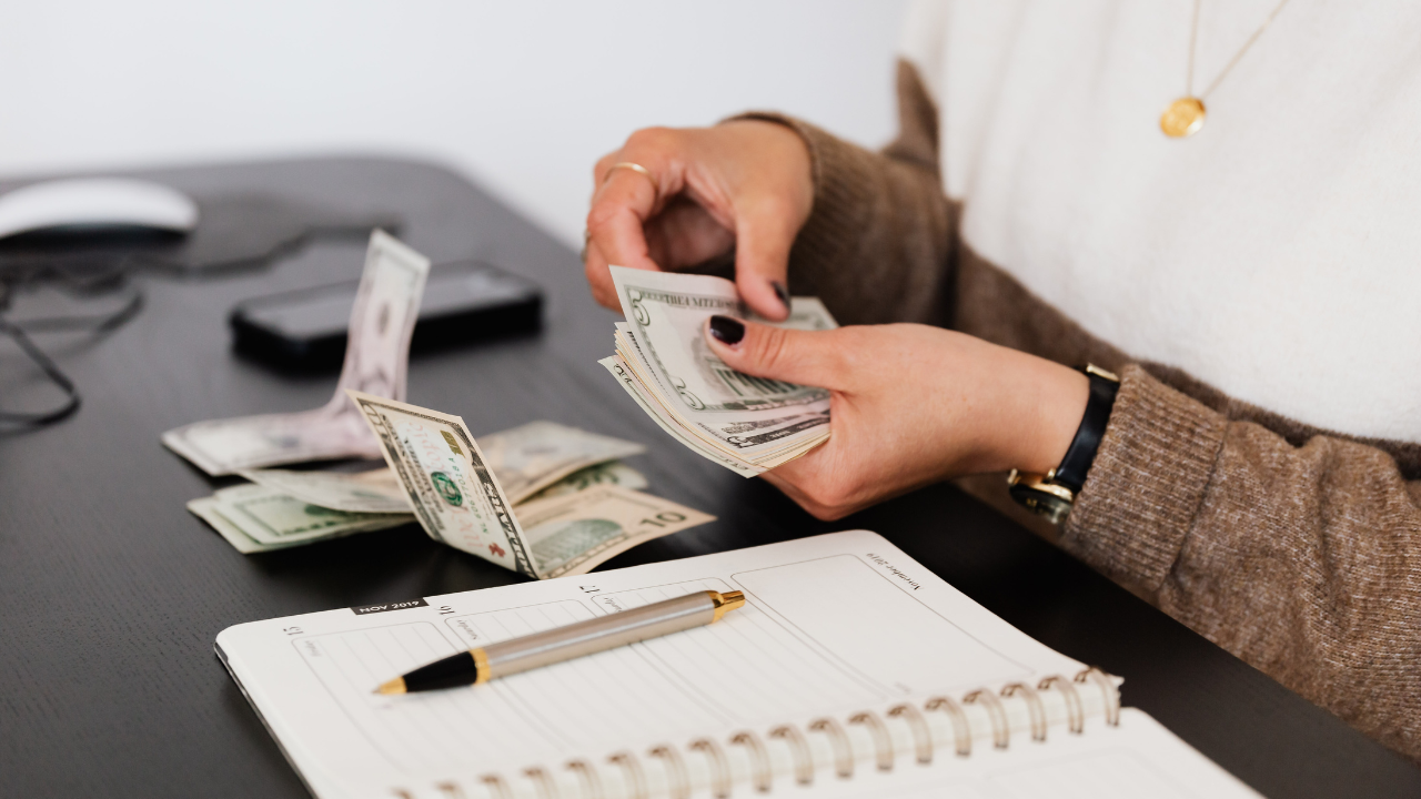 Woman counting cash at a desk with a planner, pen, and scattered bills nearby